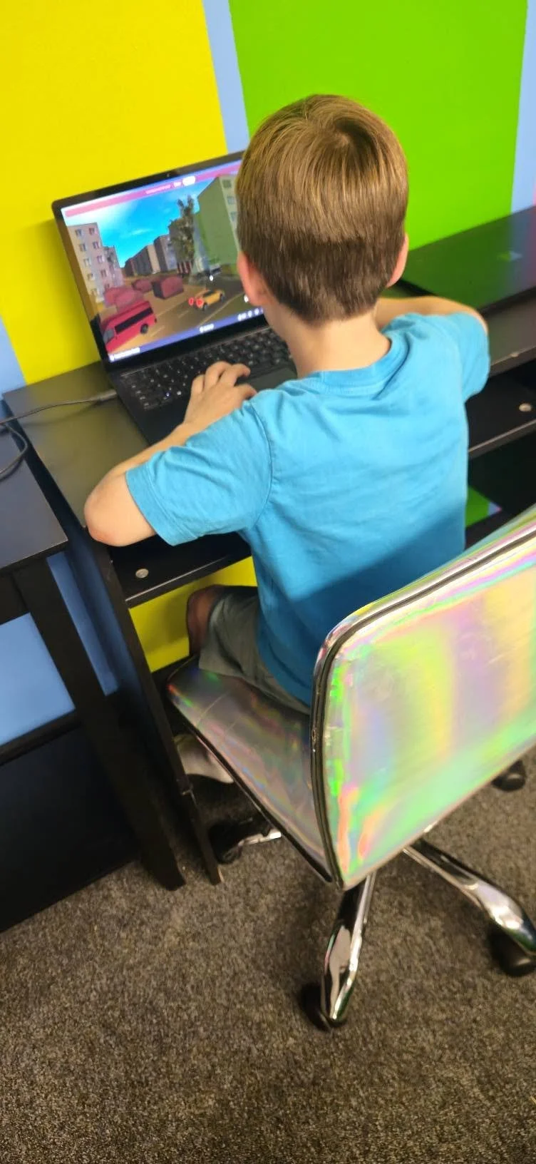 A young boy sitting at a black desk using a laptop in a colorful room with yellow and green walls.