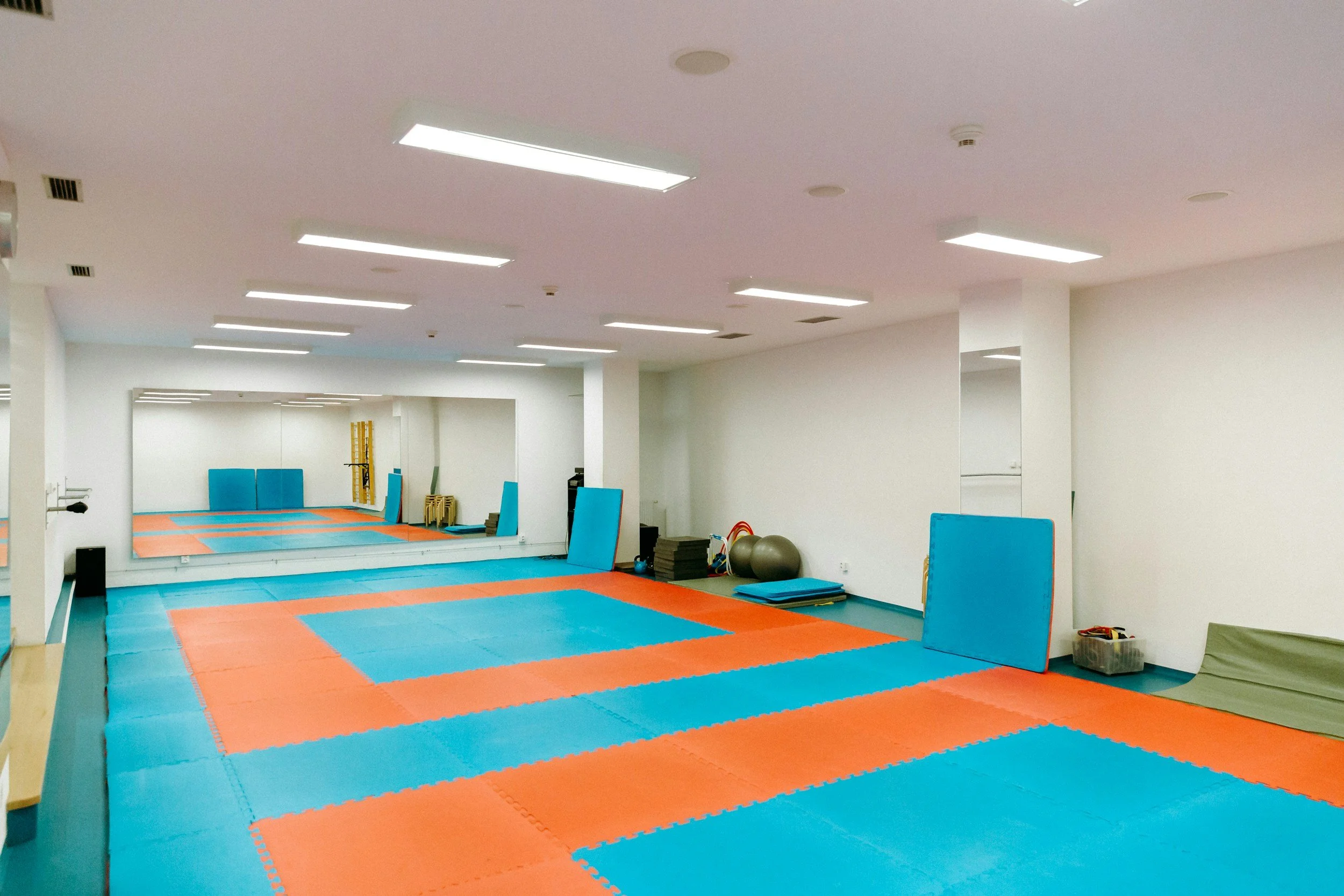 A martial arts training room with colorful interlocking foam mats, a large mirror on the wall, and exercise equipment such as mats, balls, and small weights