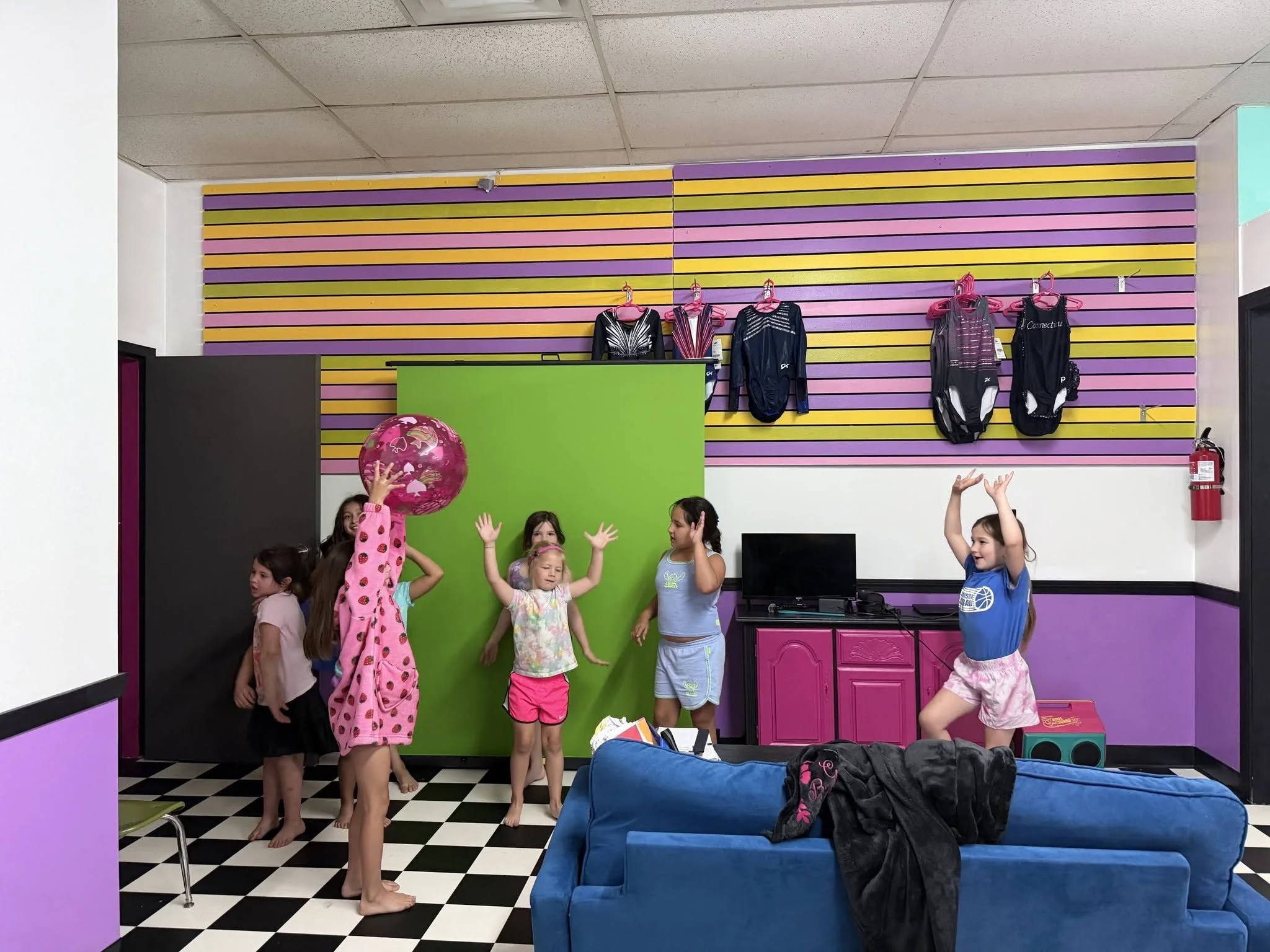 Children in pajamas and a blue shirt playing with a pink ball in a colorful room with a green partition, striped purple, yellow, pink wall, black and white checkered floor, and pink cabinets. Some children are raising their hands.