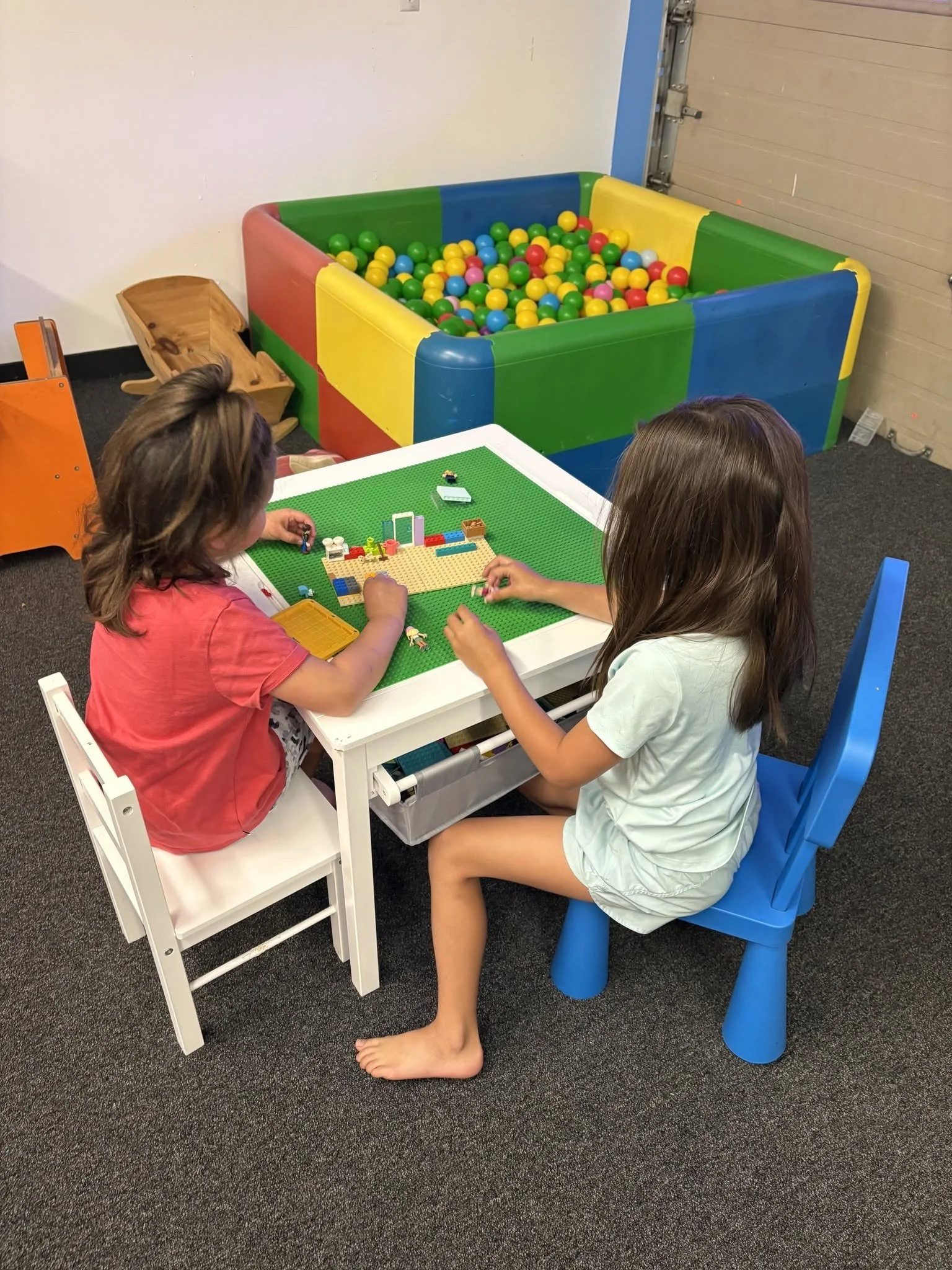 Two children sitting and playing with Lego blocks at a white table, with a colorful ball pit in the background.