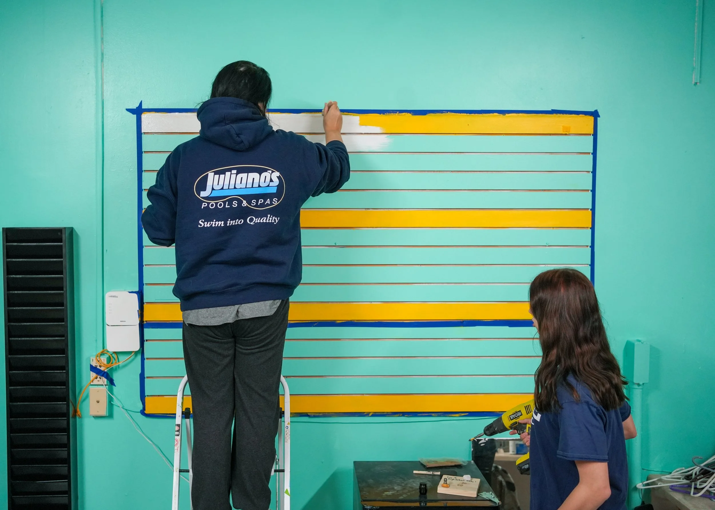 Two women installing a large blue and yellow display board on a turquoise wall. One woman, standing on a ladder, is attaching a horizontal yellow strip at the top. The other, holding a drill, is working at the bottom right.