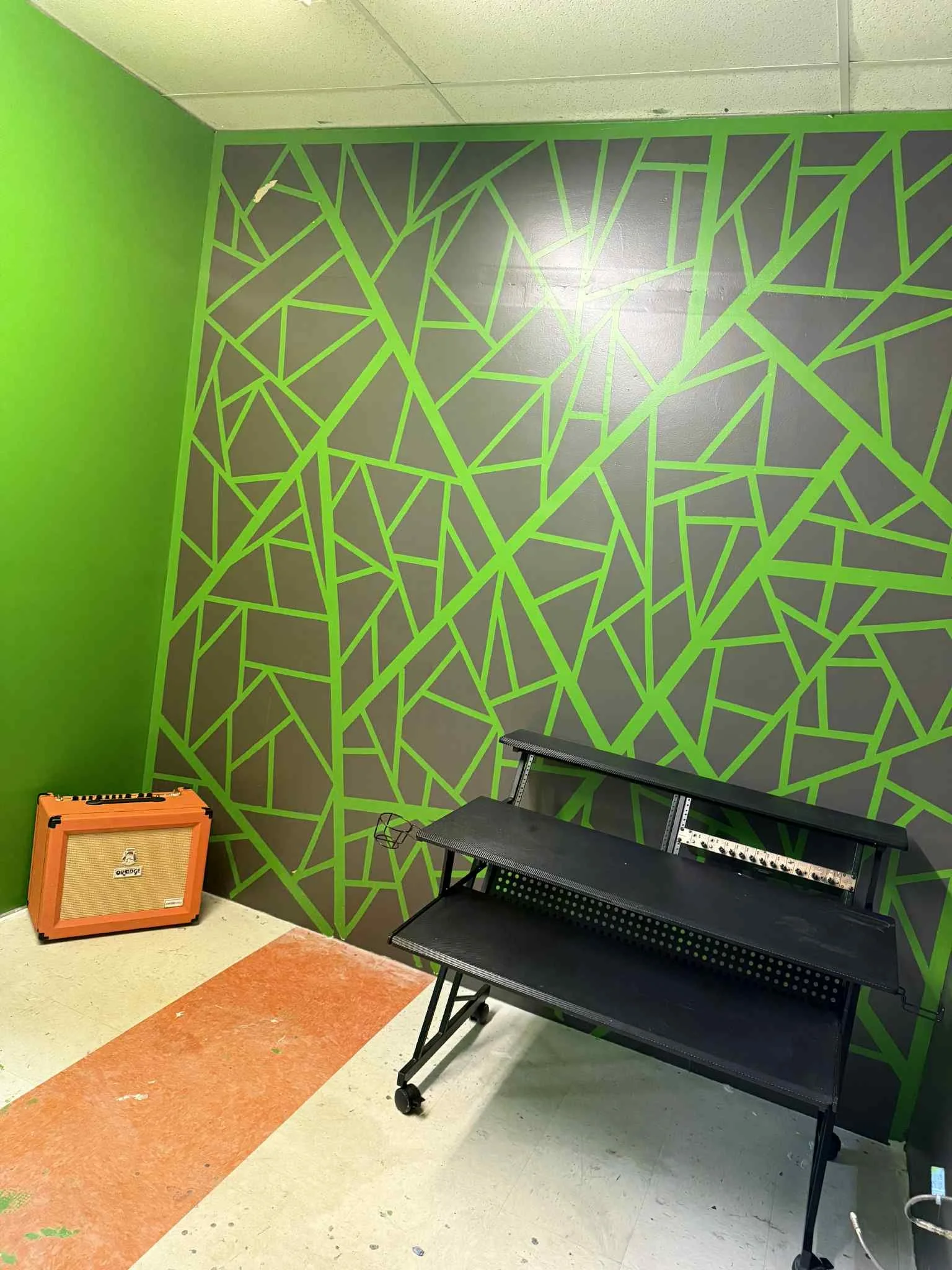 A corner of a room with a vibrant green and dark gray wall featuring a geometric pattern of interconnected shapes. There is an orange amplifier on the floor to the left and a black keyboard stand without a keyboard on it to the right.