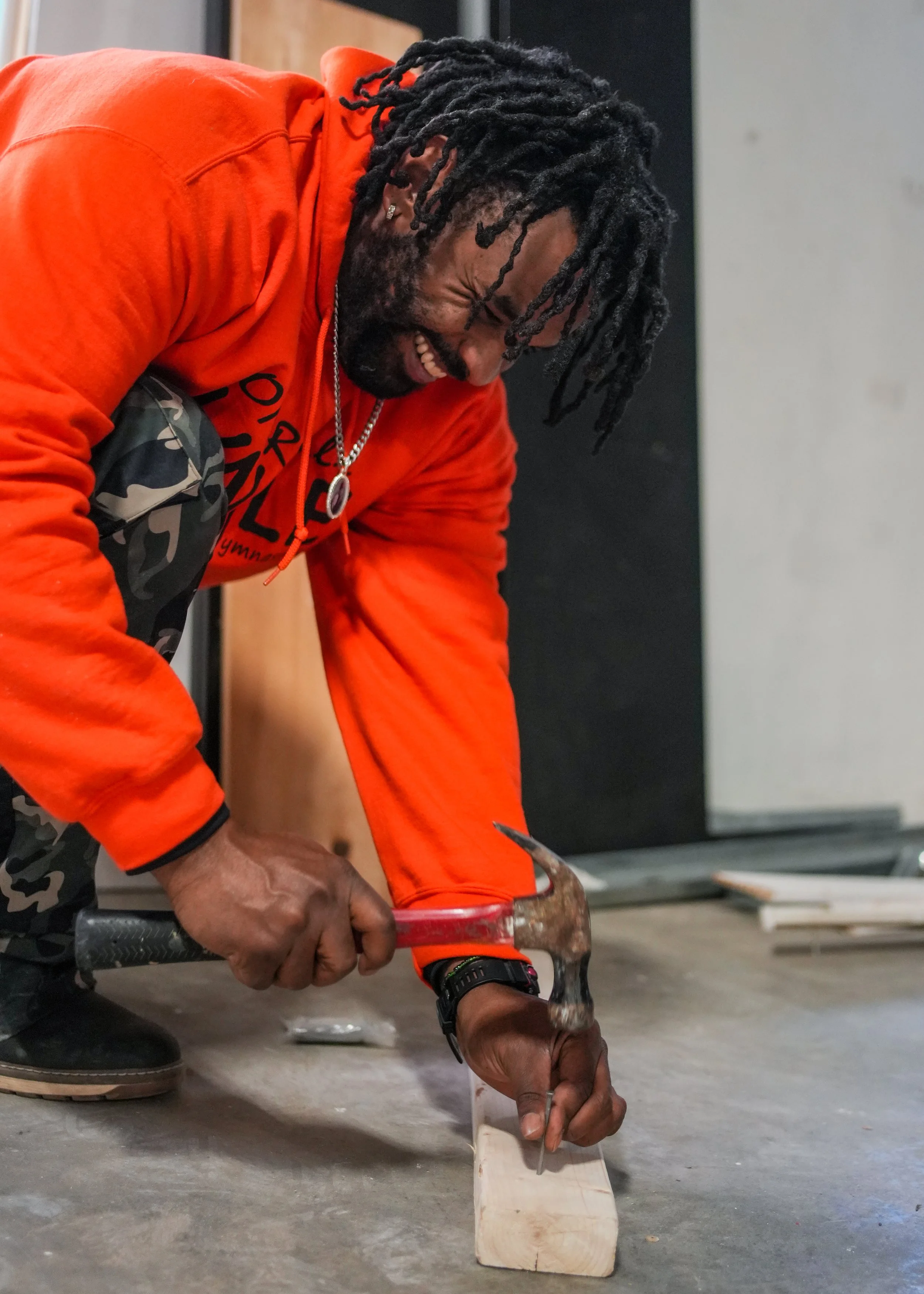 Man with dreadlocks, wearing an orange sweatshirt, using a hammer and nail to secure a piece of wood on the concrete floor.