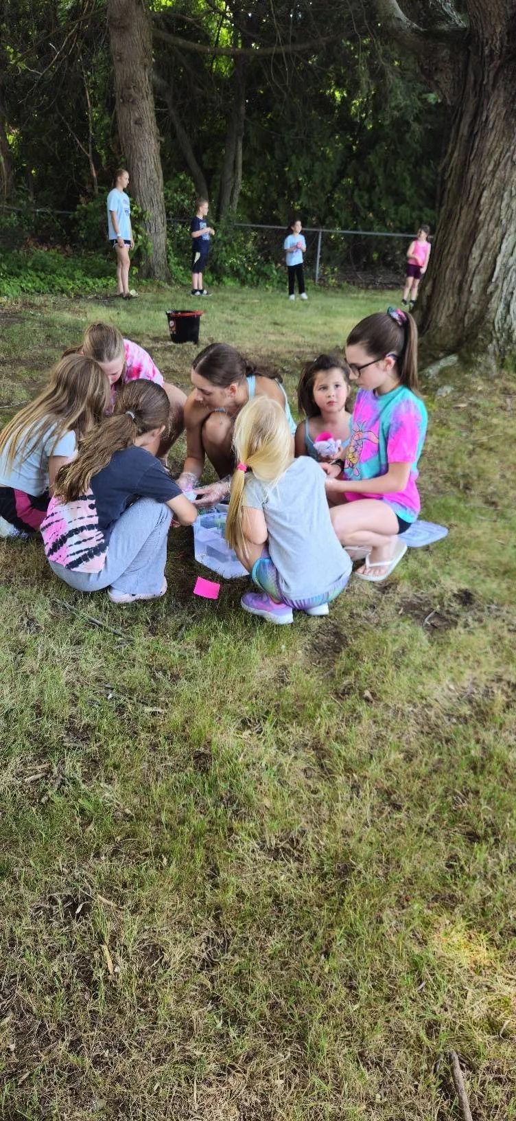 A group of children and teenagers gathered on the grass outdoors, some sitting and some standing, with trees in the background. Several children are engaging in an activity on the ground, while others are standing near a fence.