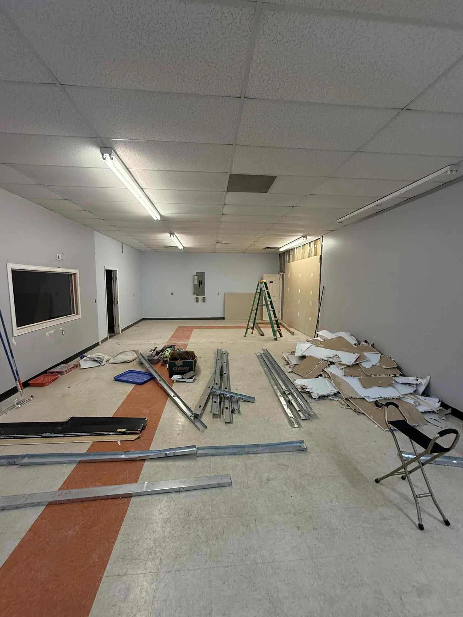 A room under construction or renovation with scattered metal framing, drywall pieces, tools, a stepladder, a folding chair, and construction debris.