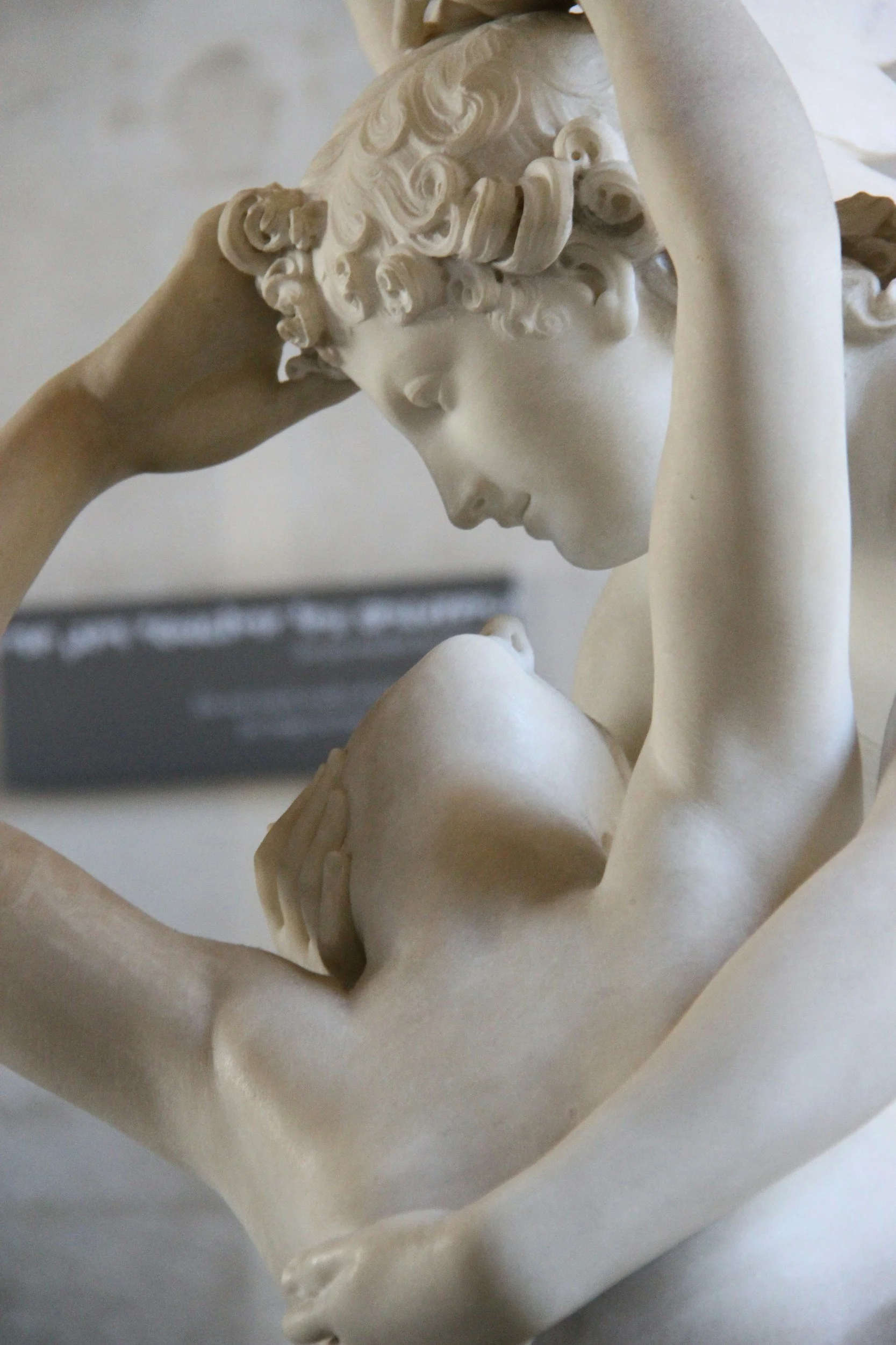 A close-up of a marble sculpture showing two figures, one with curly hair leaning down towards the other with eyes closed, and arms raised to cradle the other's head, in an intimate pose.
