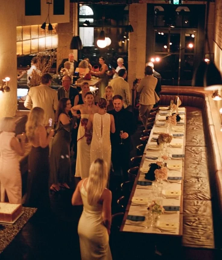 People dressed elegantly at a dinner party in a restaurant with a long table set for a meal, dim lighting, and decorative centerpieces.