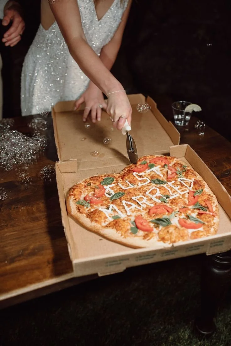 A person in a white, sparkly dress is cutting a heart-shaped pizza with the words 'US MARRIED' written on it, surrounded by a wooden table and some bubbles and a glass with a lemon slice.