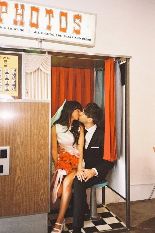 A couple sharing a kiss inside a photo booth with red and gray curtains, with a sign that reads 'PHOTOS' above them.