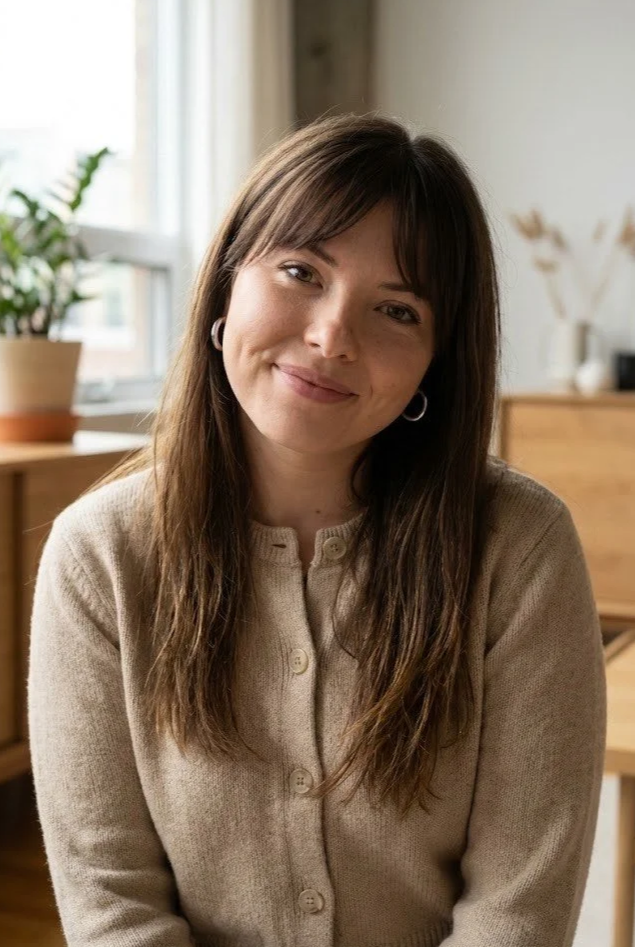 A woman with long brown hair, wearing a beige cardigan, sitting indoors near a window with potted plant and decorative items in the background.