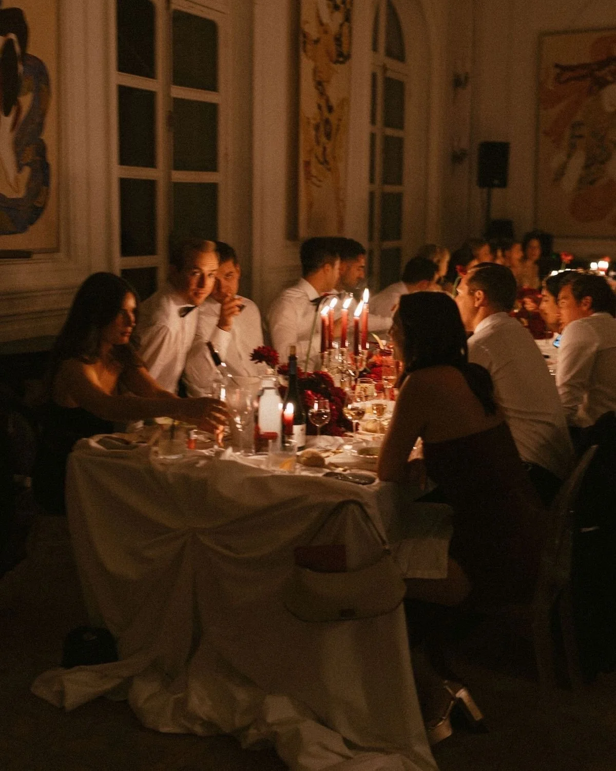 People dressed in formal attire seated around a dining table with lit candles in a dimly lit, elegant room decorated with large paintings on the walls.