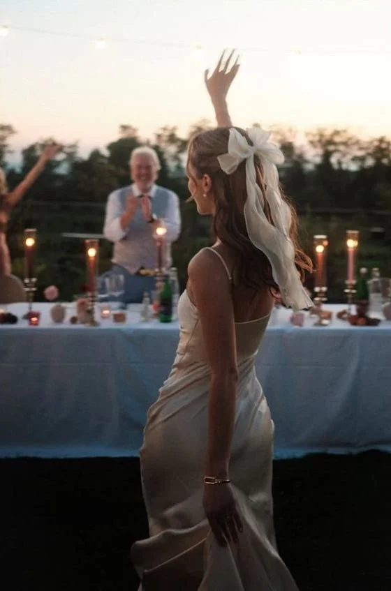 A woman with long, curly hair wearing a cream-colored satin dress and a large ribbon in her hair dancing at an outdoor evening event, with a man clapping behind a decorated table, and candles on the table creating a warm ambiance.