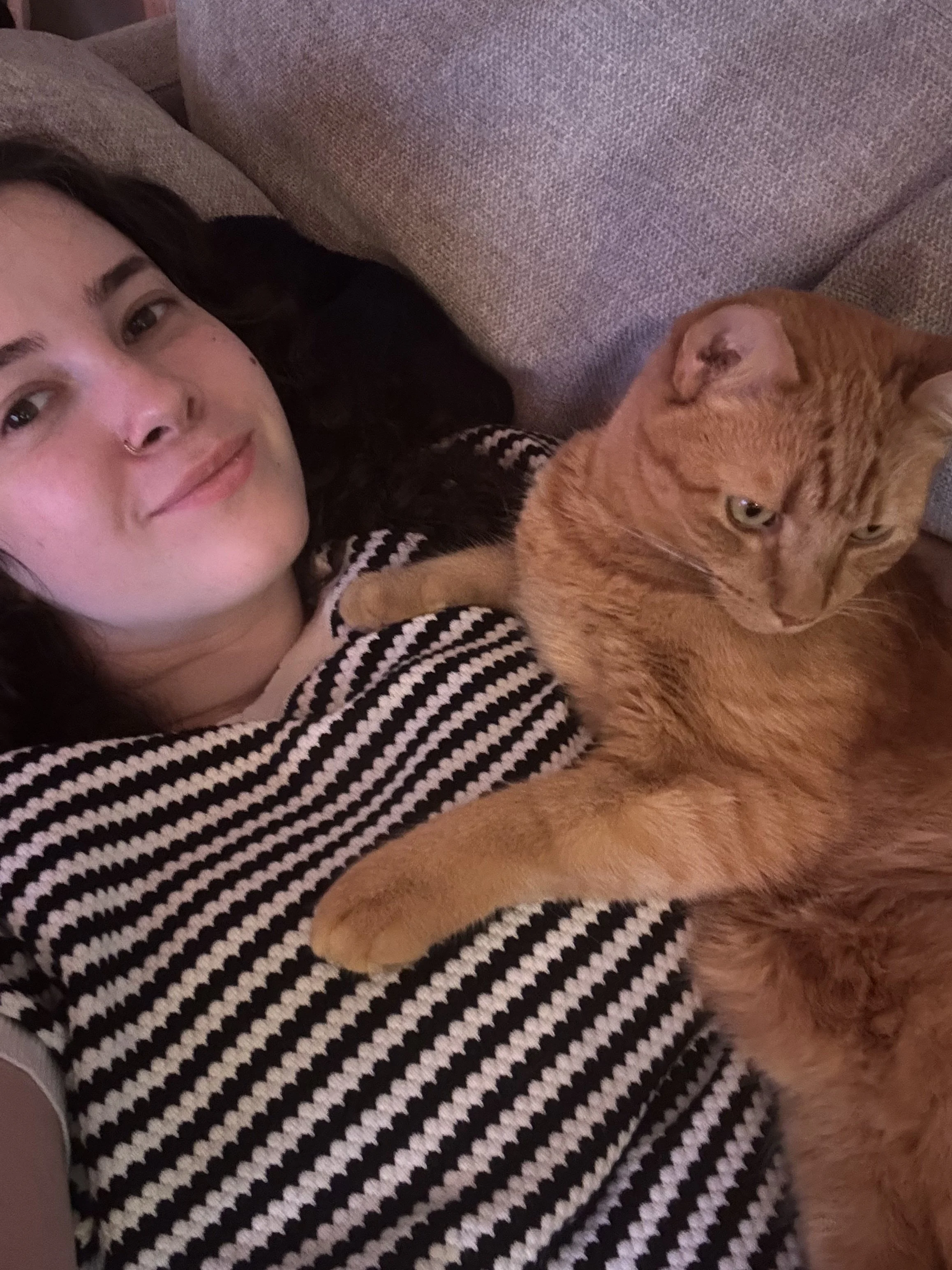 A woman lying on a brown couch next to an orange tabby cat.