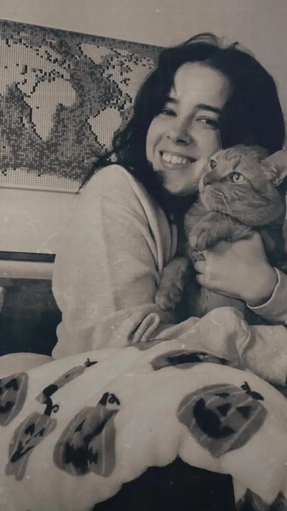 A woman smiling and holding an orange tabby cat, indoors with a mosaic wall art in the background, in black and white.