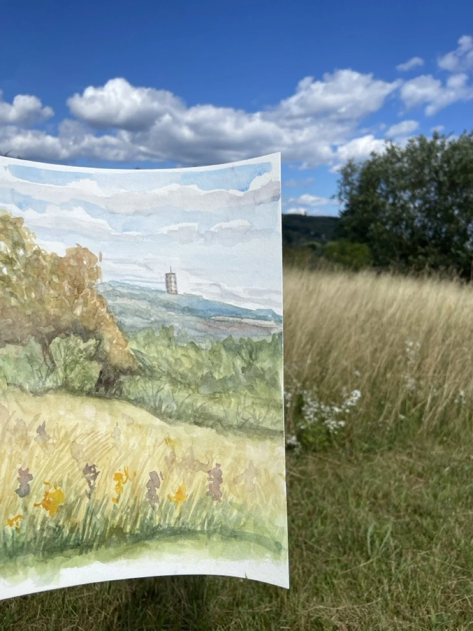 Watercolor painting of a Duluth, Mn’s Enger Tower with trees, a tower in the distance, grassy fields, and a cloudy sky, held up outdoors with natural surroundings in the background.