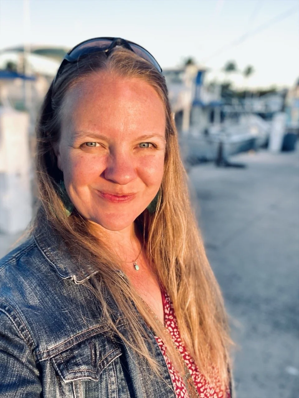 Anni Friesen has  long blond hair, and is wearing sunglasses on her head, a denim jacket, and a red patterned top, smiling outdoors near a marina with boats in the background, during sunset.