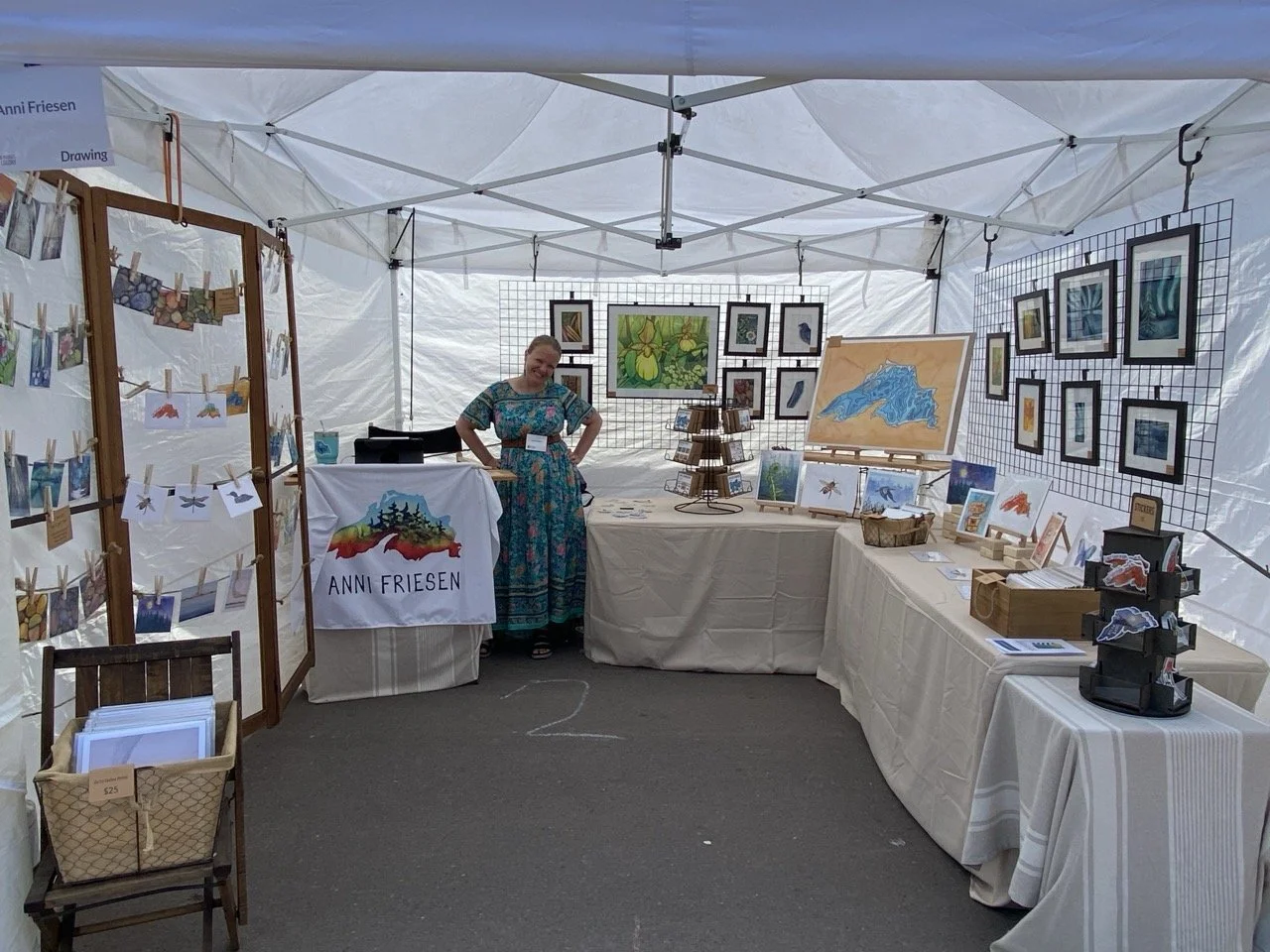 Art booth at outdoor market with paintings, photographs, and crafts, featuring a woman in a colorful dress standing behind a table with a white cloth that says 'Anni Friesen'.