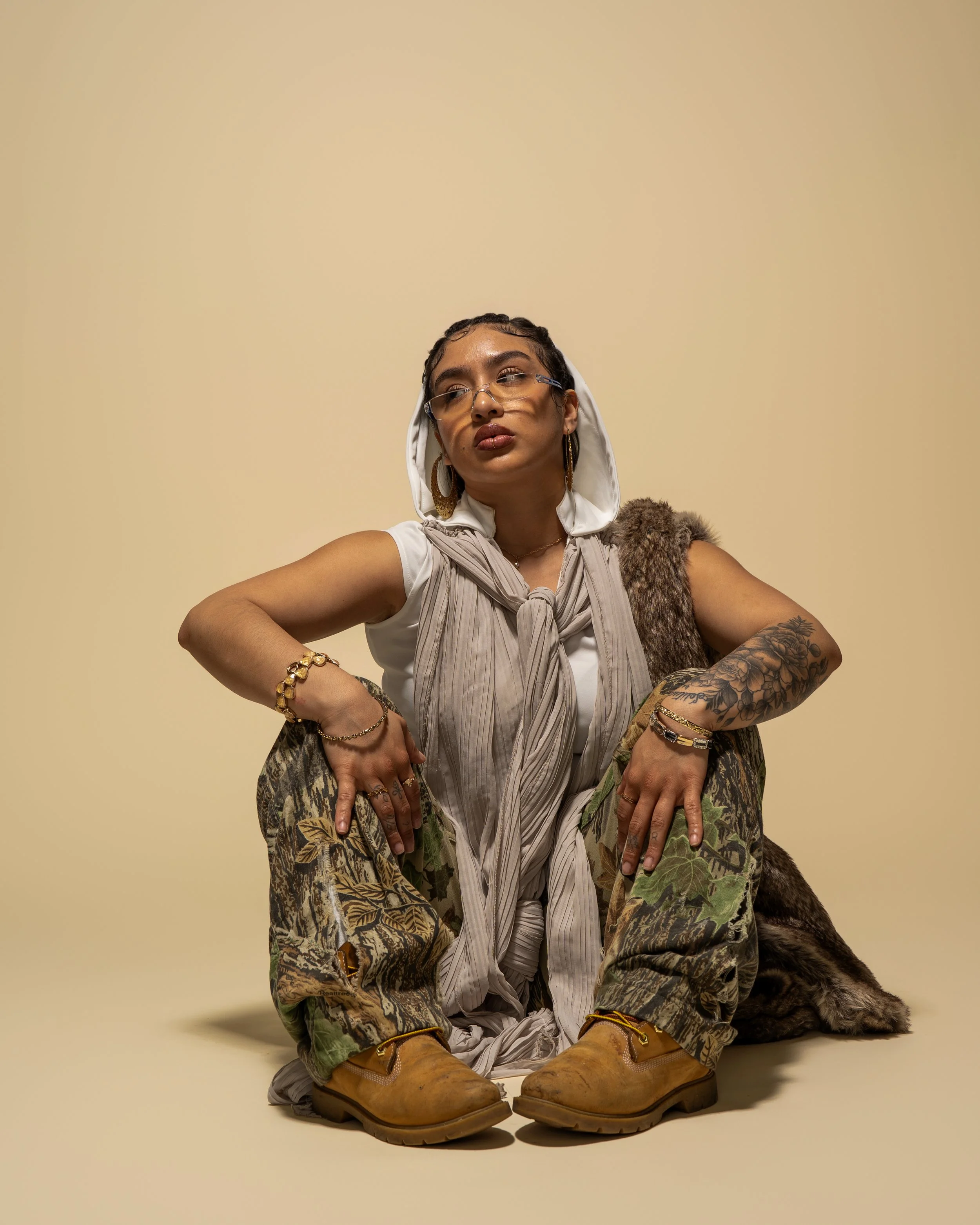 Person sitting on the ground in a stylish outfit, wearing camouflage pants, tan boots, layered jewelry, glasses, and a hooded vest with a furry coat draped over her back, posing against a plain beige background.