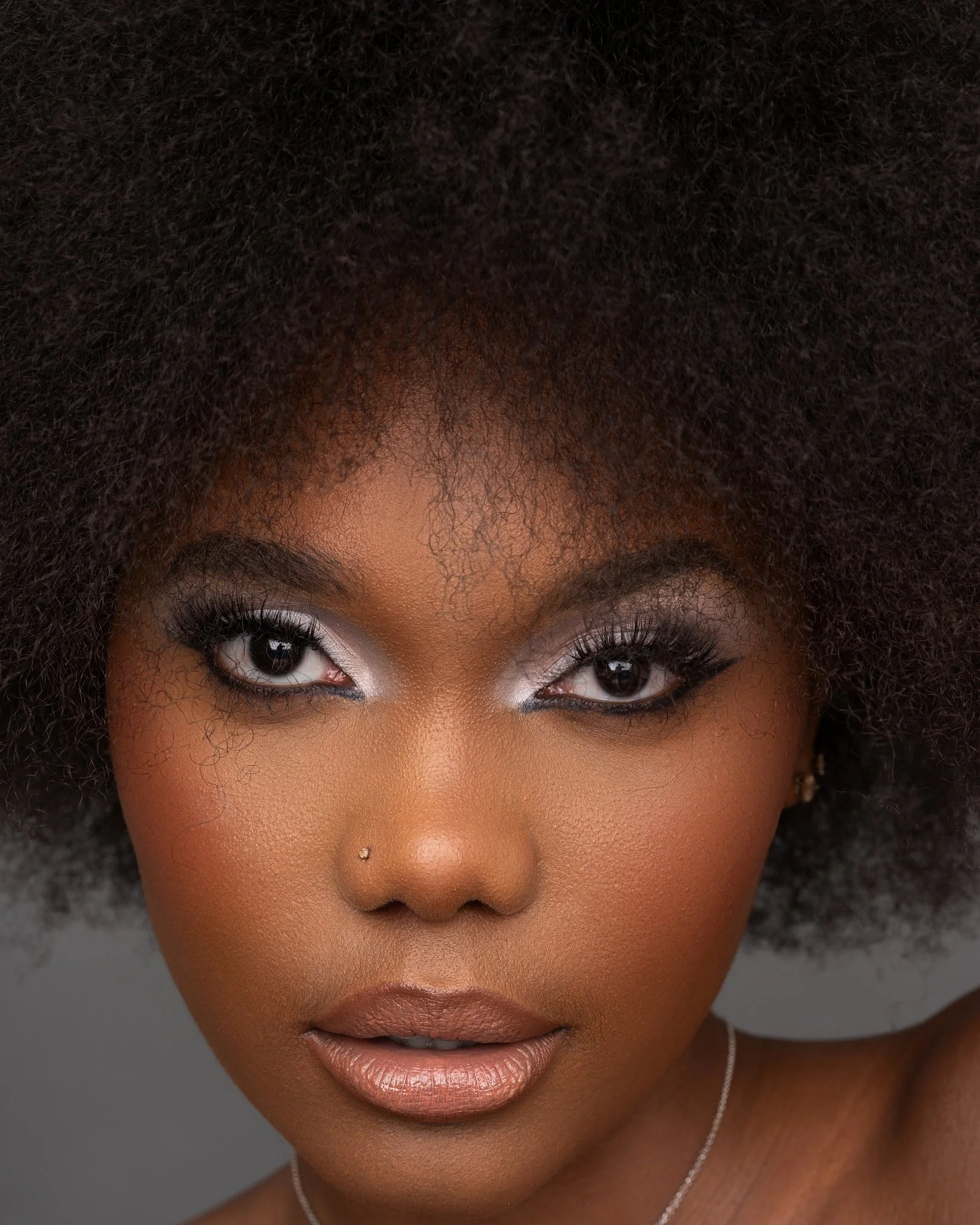 A close-up portrait of a woman with dark skin, curly afro hairstyle, silver eye makeup, and a nose piercing, looking directly at the camera.