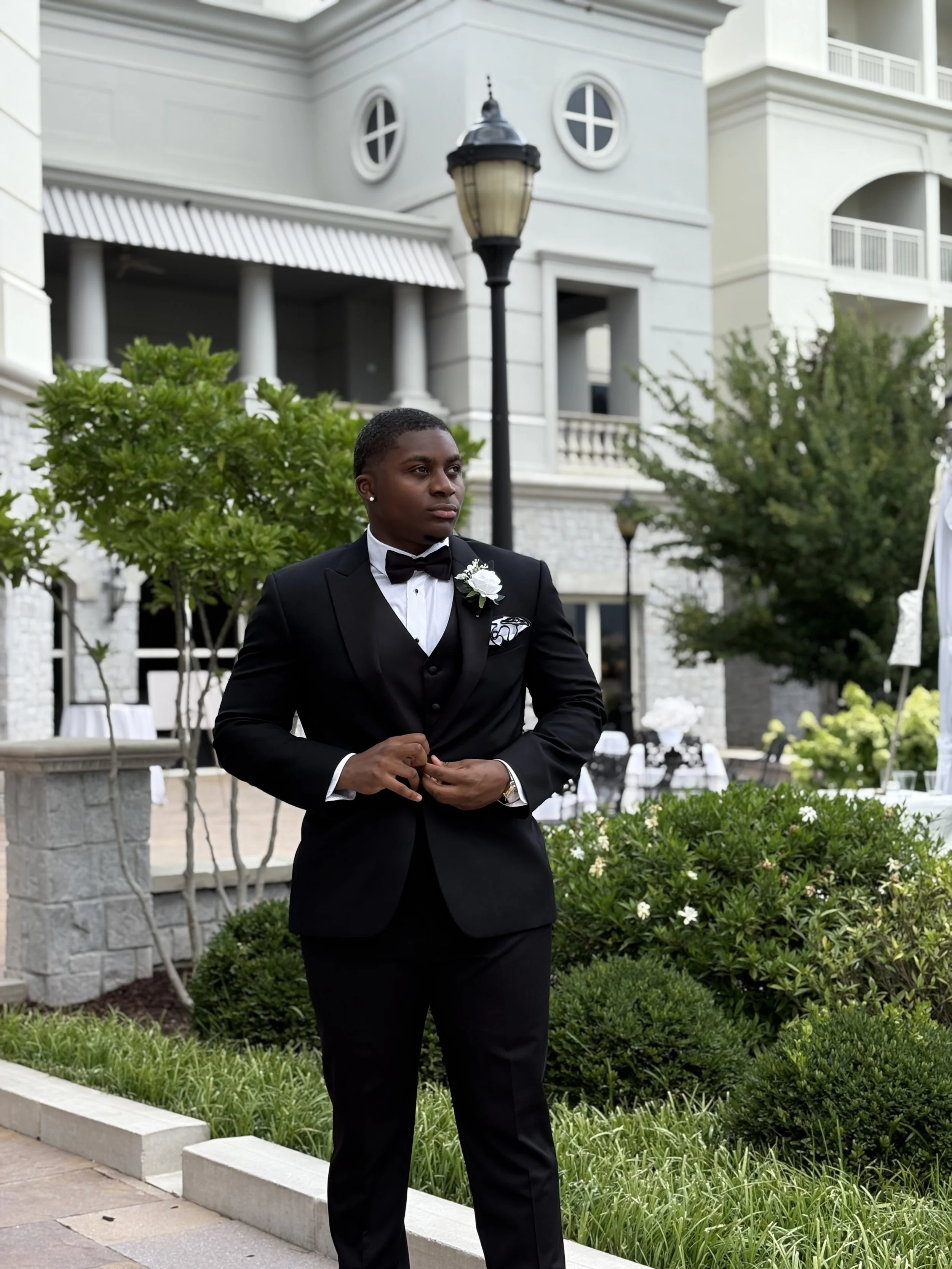 A person dressed in a black tuxedo with a bow tie, white shirt, and boutonniere, standing outdoors in a formal setting, with a building and greenery in the background.