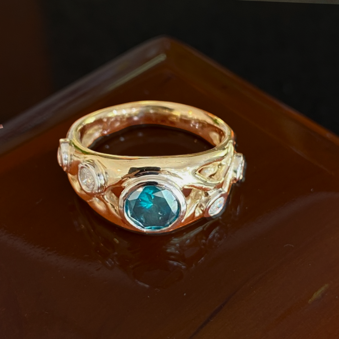 A gold ring featuring a large blue gemstone in the center, surrounded by smaller clear gemstones, placed on a dark wooden surface.