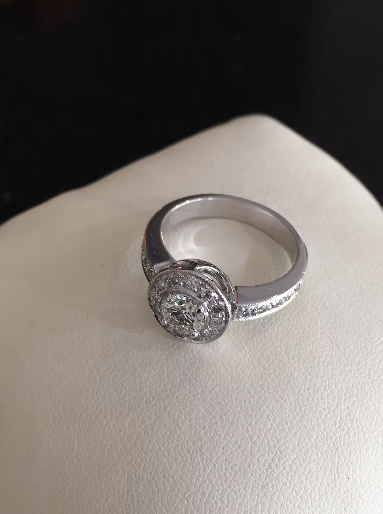 A silver ring with a large round gemstone surrounded by smaller gemstones, displayed on a white surface.