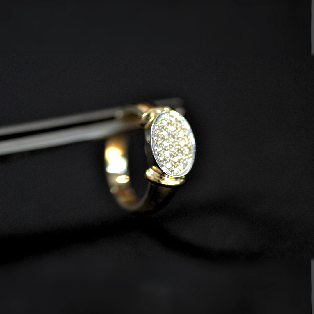A gold ring with a large oval face encrusted with small diamonds, hanging from a pair of tweezers against a black background.