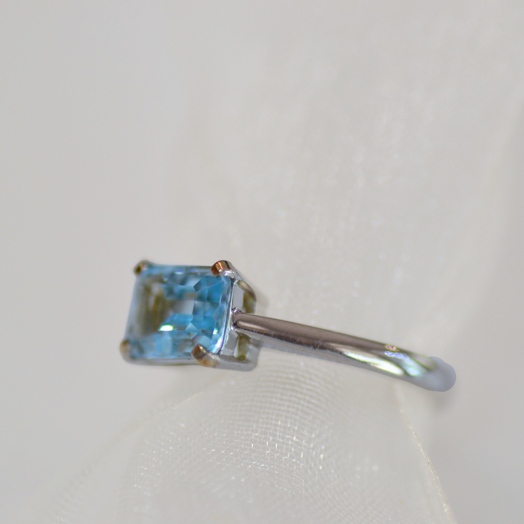 A silver ring with a rectangular blue gemstone, photographed close-up on a white textured surface.