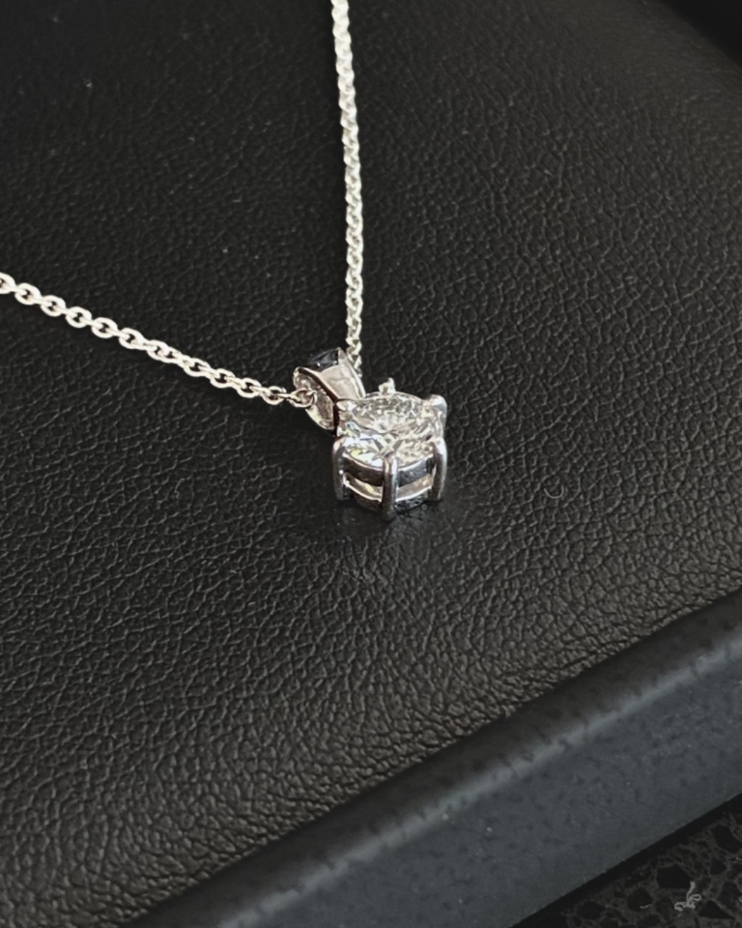 A close-up of a silver necklace with a princess-cut diamond pendant placed on a black textured surface.