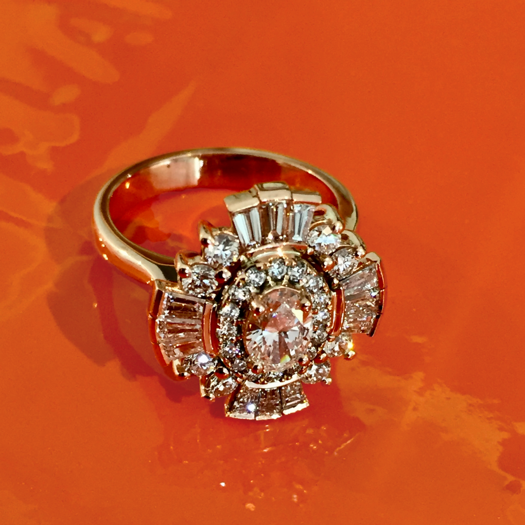 A close-up of a decorative ring with a large central gemstone surrounded by smaller diamonds, set on an orange surface.