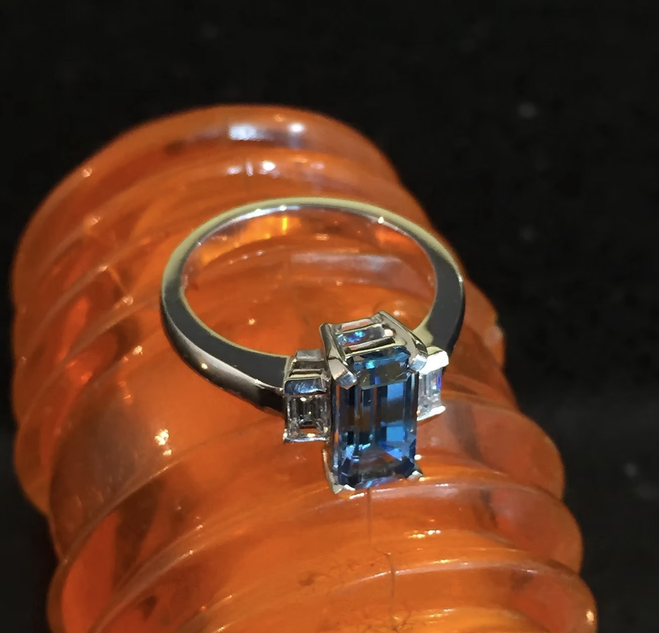 Silver ring with a large rectangular blue gemstone, flanked by two smaller clear stones, placed on an orange spiral object against a black background.