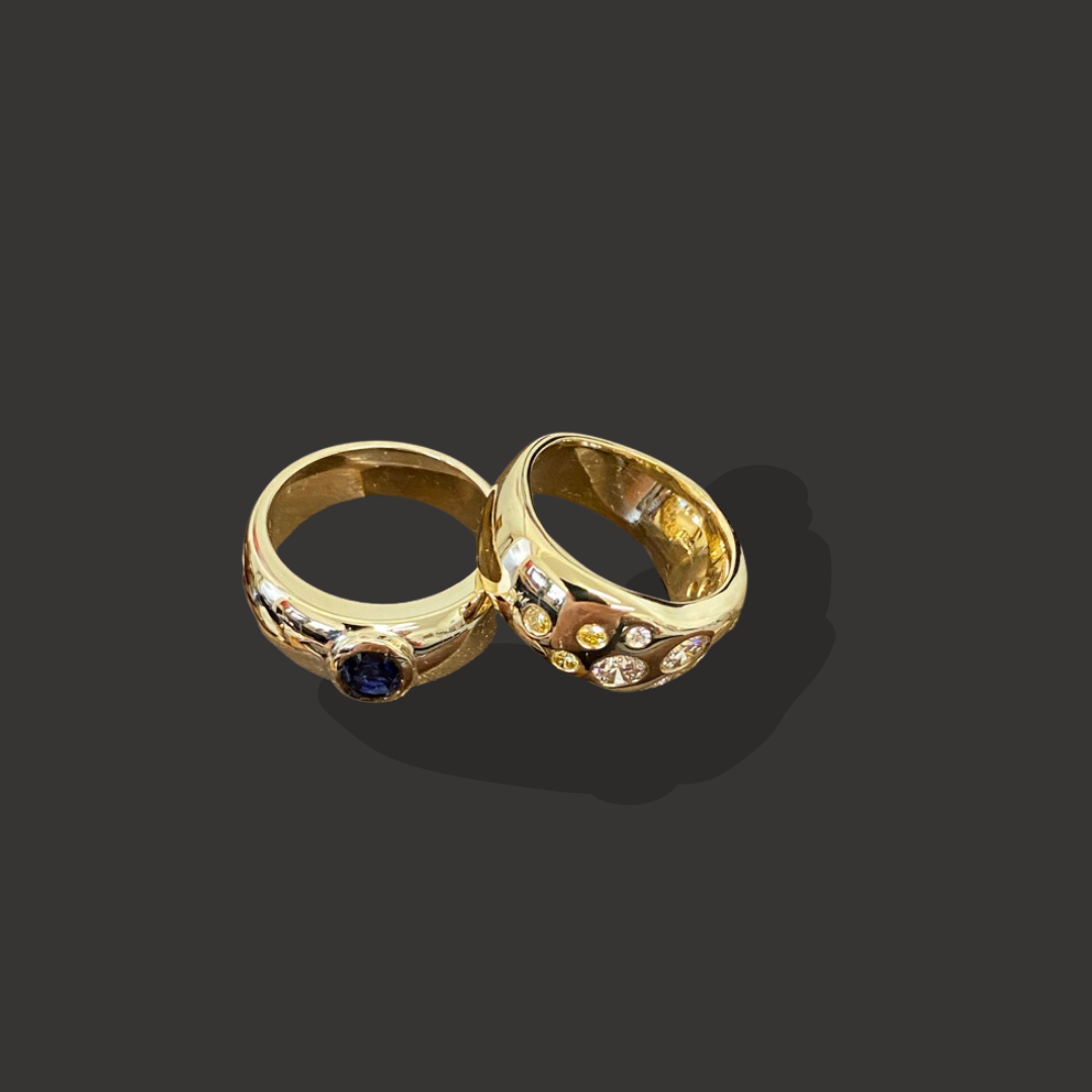Two gold rings, one with a blue gemstone and the other with multicolored diamonds, on a dark background.