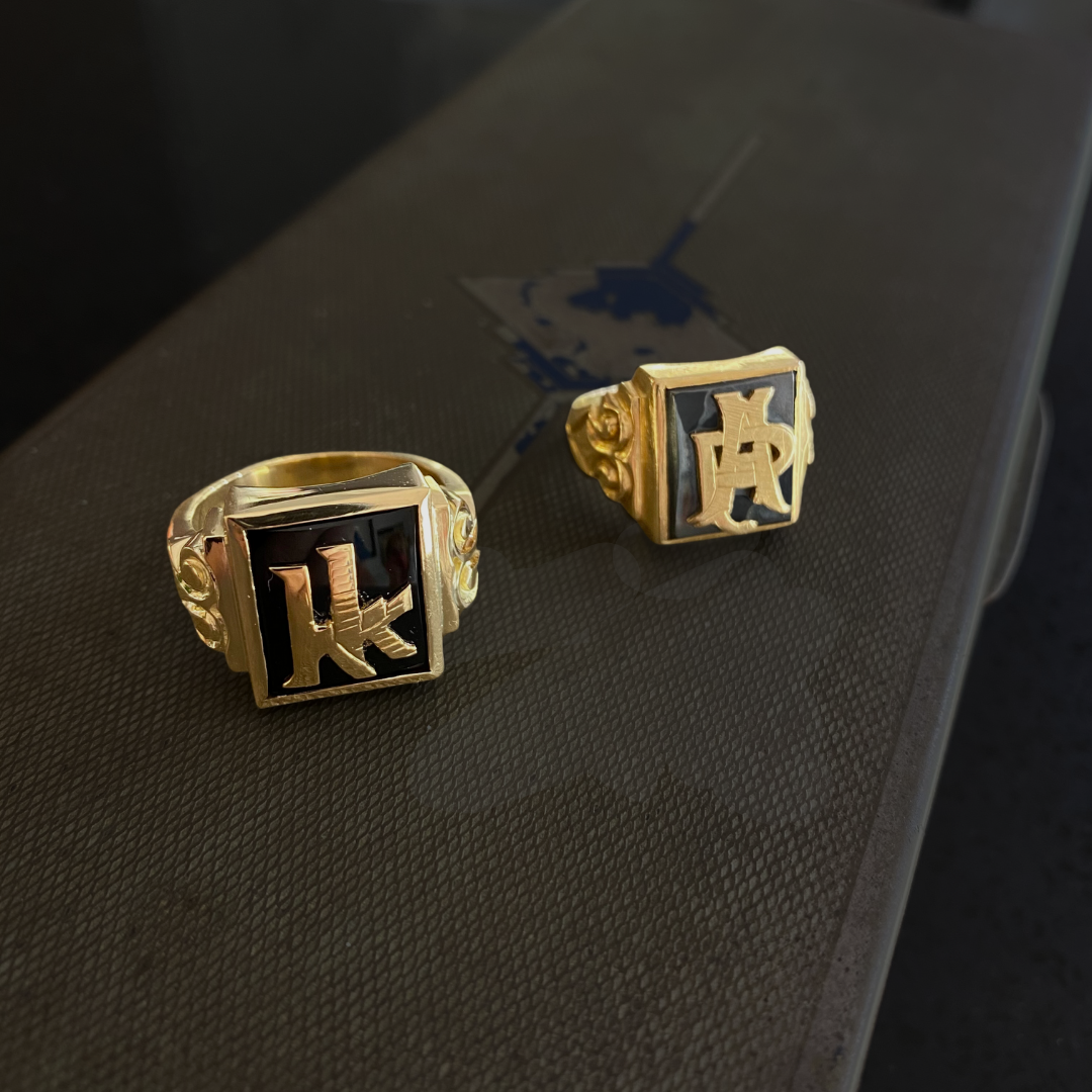 Gold rings with black background and gold 'K' and 'A' etched on the top, placed on a textured black surface.