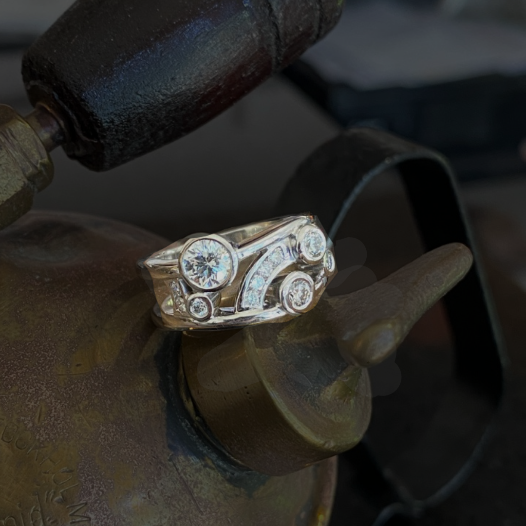 A silver ring with multiple diamonds is placed on a metal surface, with a welding tool in the background.