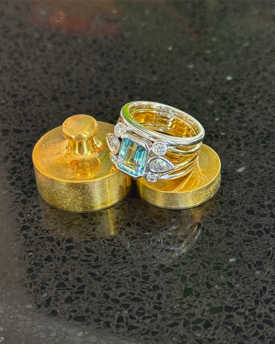 A jewelry ring with a large rectangular blue gemstone surrounded by smaller diamonds, resting on two gold-colored weighing scales on a black textured surface.