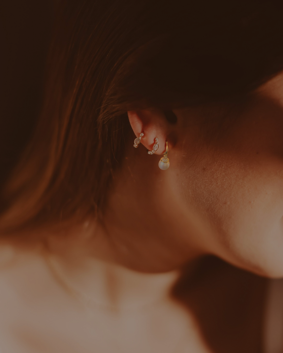 Close-up of a woman's ear with multiple earrings, including a pearl drop earring, with brown hair framing her ear.