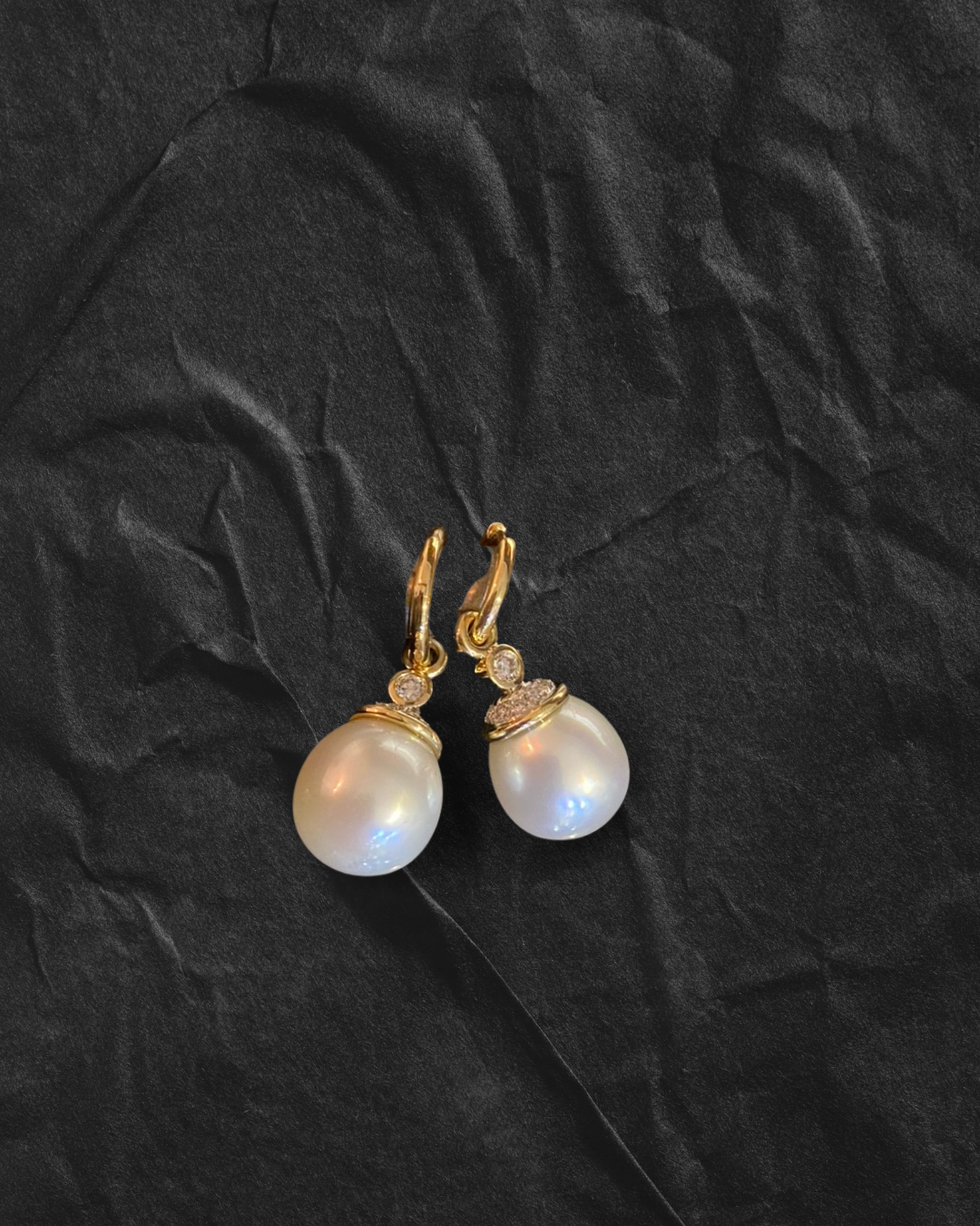Pair of pearl drop earrings with gold hooks and small diamonds on a black textured background.