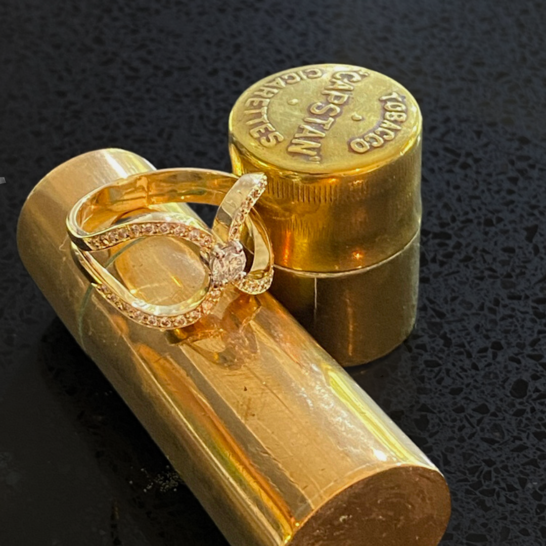 A gold lipstick tube, a gold container with embossed text, and a gold ring with a large center stone surrounded by smaller stones, placed on a black textured surface.