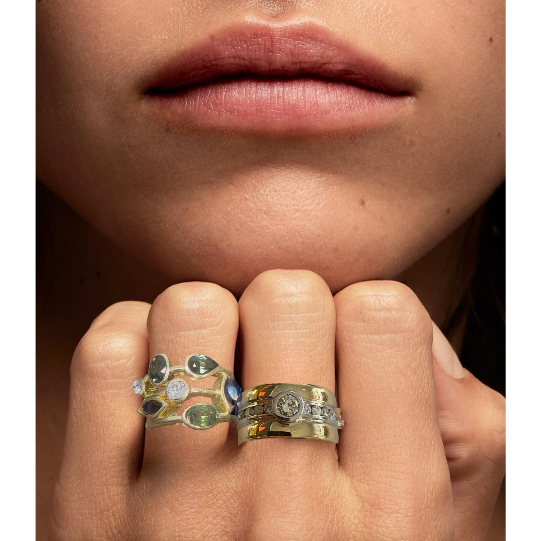 Close-up of a person's lips and hand, showing two gold rings with gemstones and diamonds on their fingers.