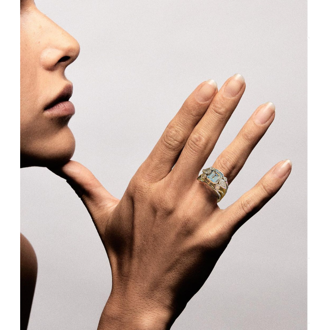 A close-up of a woman's face, hand, and fingers. She is wearing an ornate ring with a large square gemstone and smaller stones around it on her ring finger.