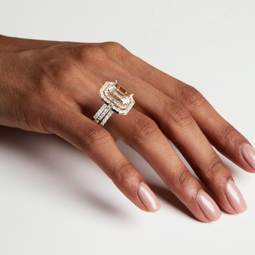 Close-up of a hand wearing an ornate diamond and gold ring with a large rectangular gemstone, resting on a white surface.