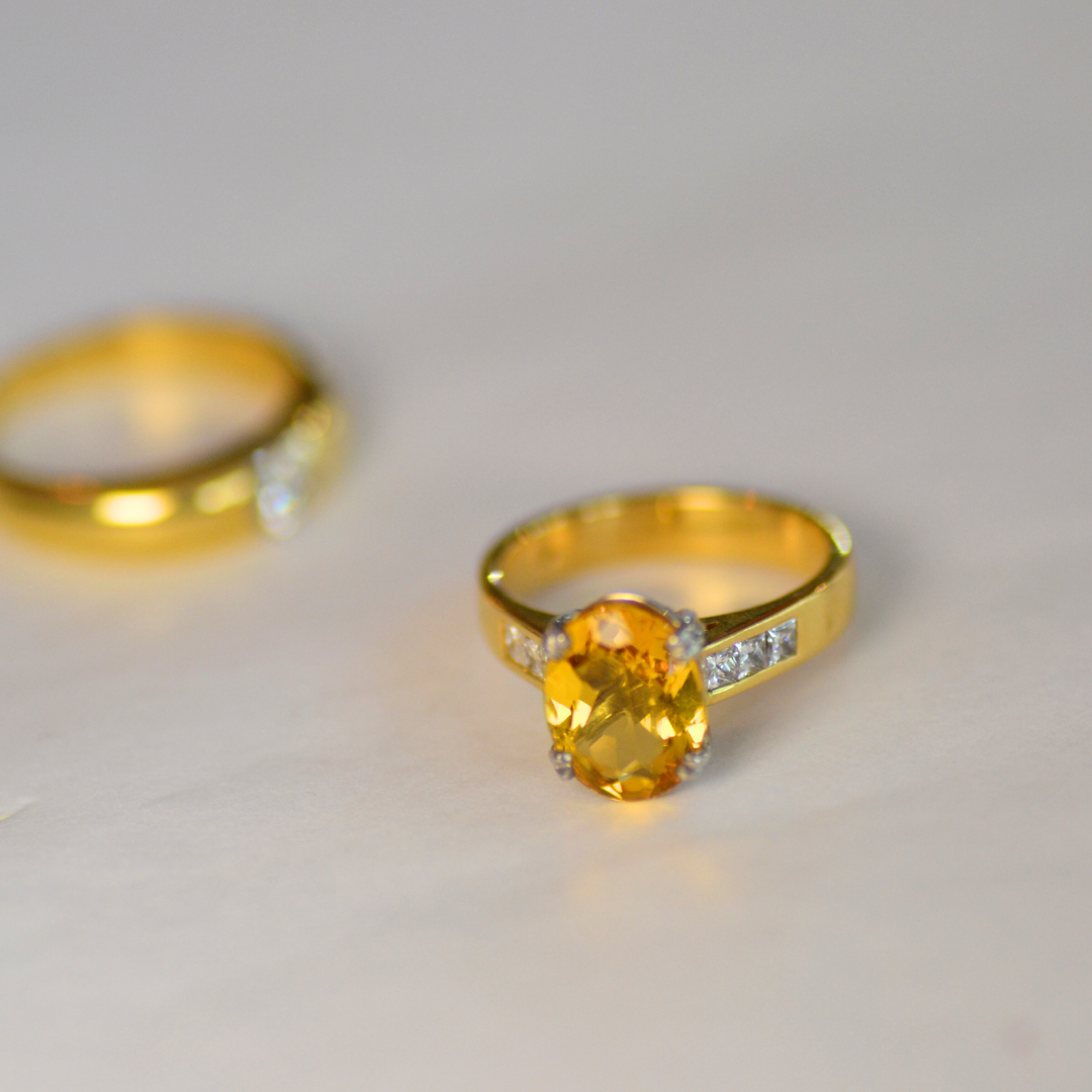 Gold ring with a large yellow gemstone and small white gemstones on the band, placed on a white surface with a blurred gold ring in the background.