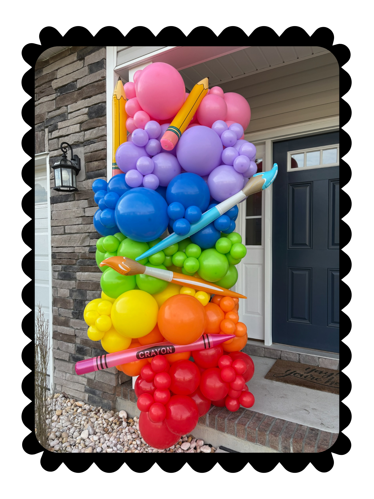 Colorful rainbow balloon column with oversized pencils, paintbrushes, and crayons displayed at a front door for a creative themed event.