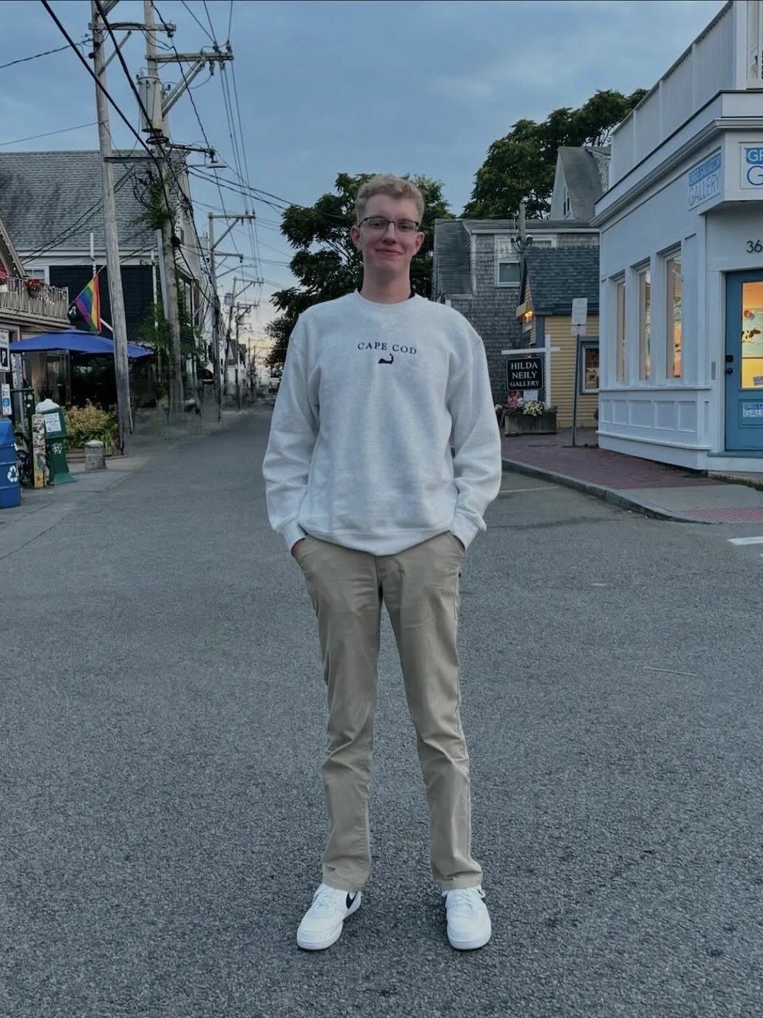 Young man standing on a street in a small town at dusk, wearing glasses, a white sweatshirt with 'Cape Cod' and a whale graphic, beige pants, and white sneakers.