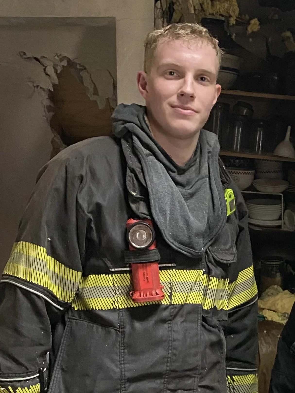 Young male firefighter in uniform, smiling, standing indoors with shelves of dishes and decor in the background.