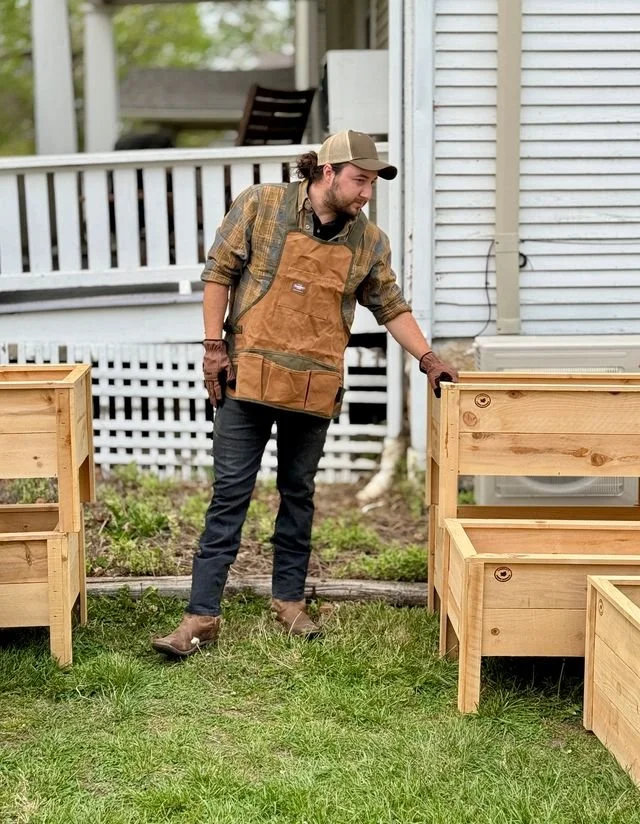 A man in a plaid shirt, jeans, and brown boots with gardening gloves and a tan cap is inspecting or building wooden garden boxes outside a house with white siding and a porch.