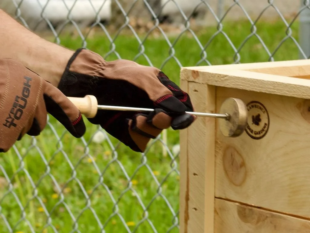 Person using a screwdriver to assemble a wooden box or piece of furniture outdoors near a chain-link fence.