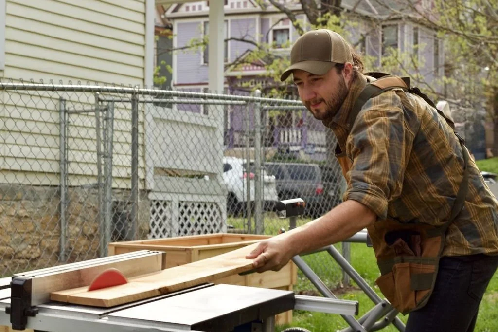 Man in plaid shirt and cap sawing a wooden piece on a workbench outdoors.