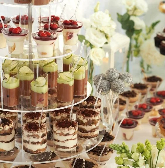 Assorted mini desserts on a tiered display, including cream cups with cherries, layered mousse in glasses, and tiramisu cups, with a side of decorated cake pops and flowers.