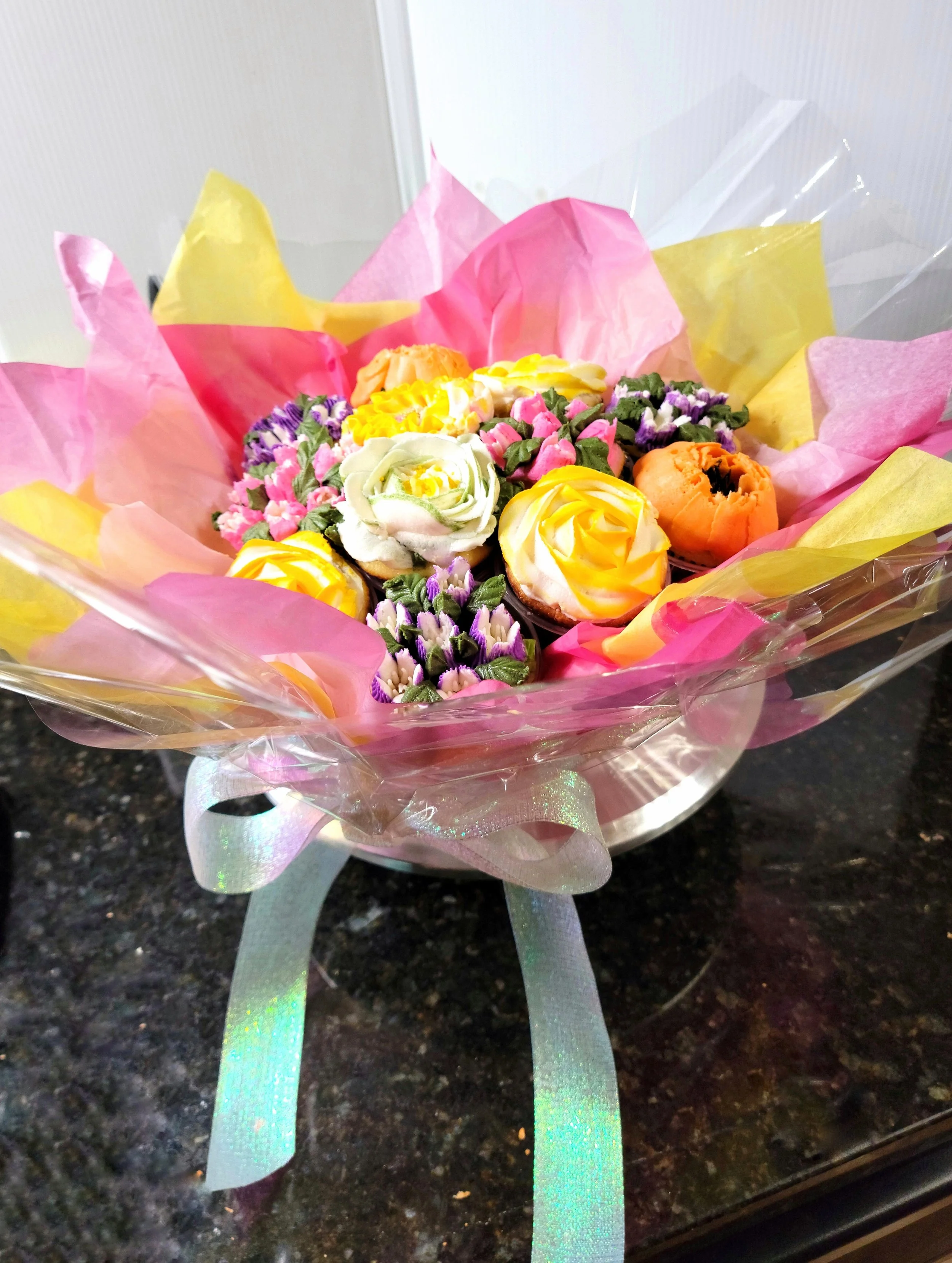 Bouquet of decorated cupcakes and flowers wrapped in pink, yellow, and clear cellophane with a ribbon, placed on a dark countertop.